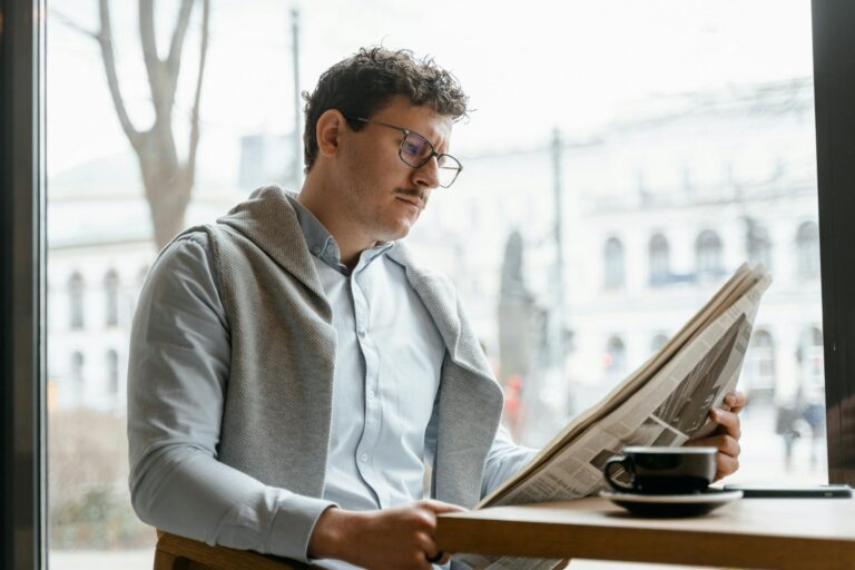 Adult man reads a newspaper by a café window, enjoying a coffee break indoors.