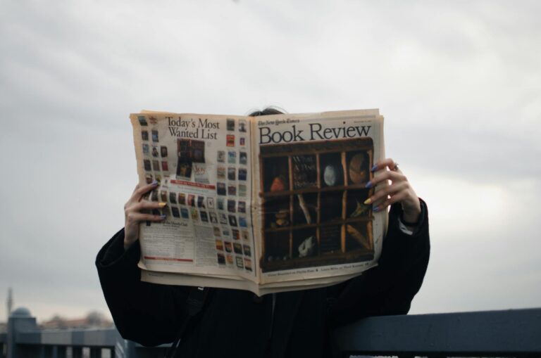 Individual holding a book review newspaper with colorful nails against a cloudy sky backdrop.